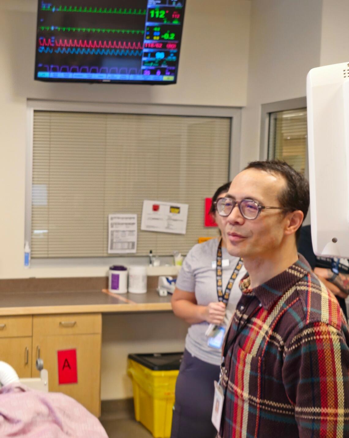 A man in a plaid shirt and glasses stands in a hospital room
