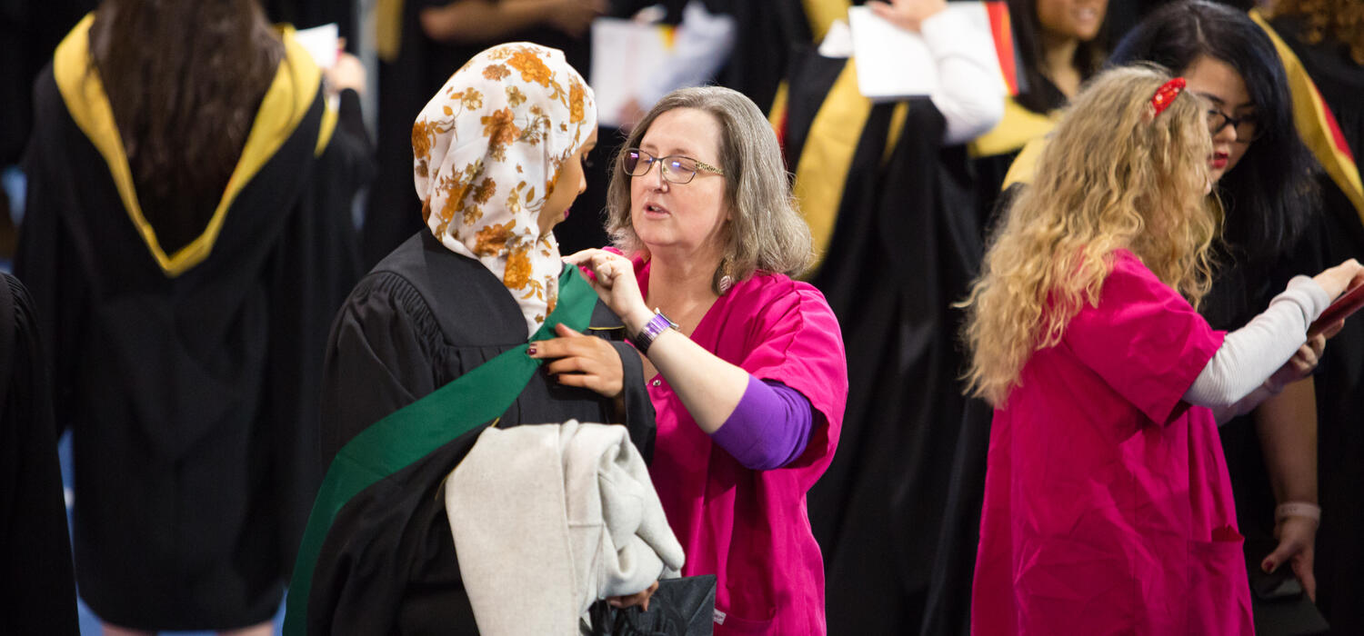 Two volunteers help graduates pin on gowns and provide directions as the grads get ready to walk the stage.