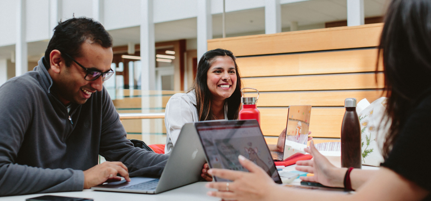 Two students laugh at a table, which has laptops on it. 