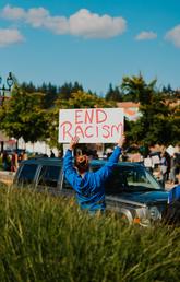 A woman stands facing traffic holding a sign that says "End Racism."