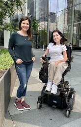 A woman in a wheelchair poses next to a woman standing with her hands in her pockets. Both women are smiling at the camera.