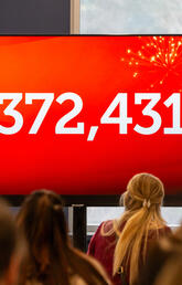 A group of people face a large red screen with '$372,431' on it