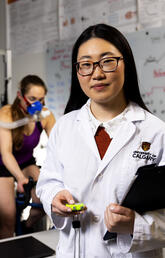 A woman wearing a lab coat and glasses smiles at the camera while another works out behind her