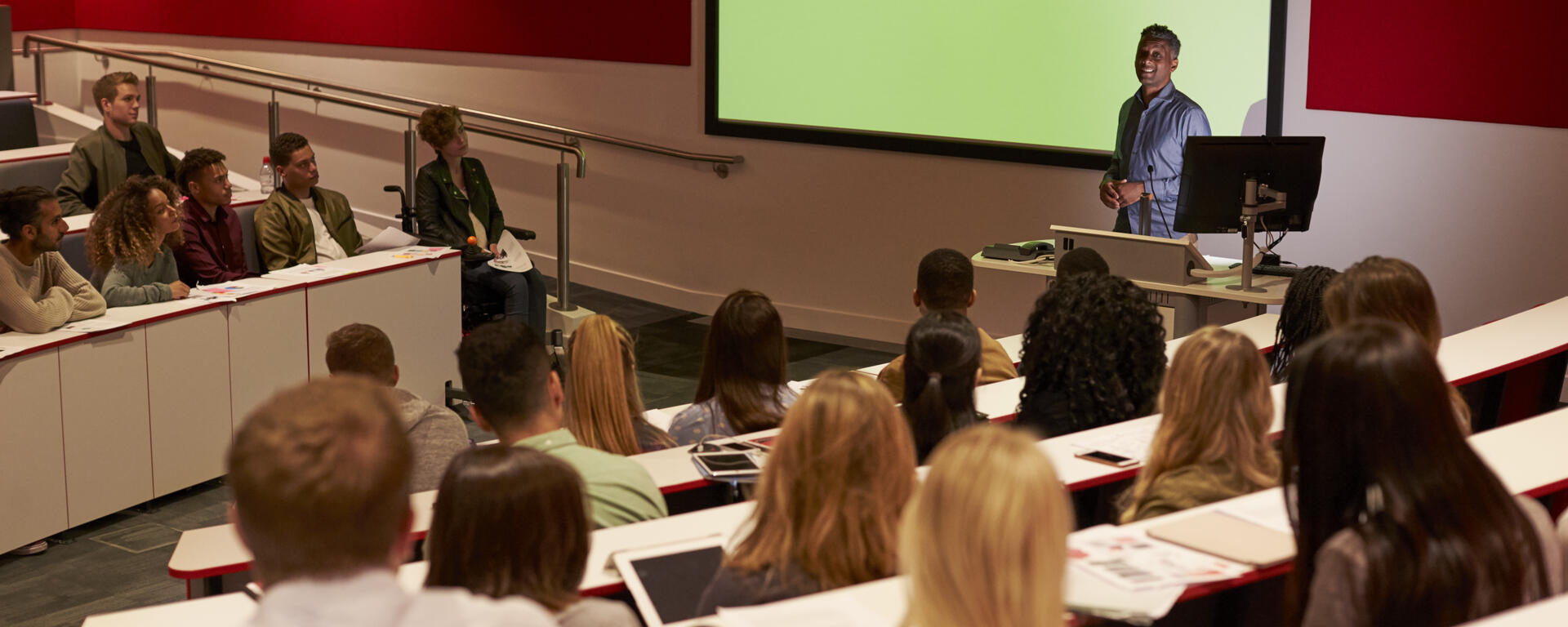 classroom with students watching and listening to a professor speak in front of large screen 